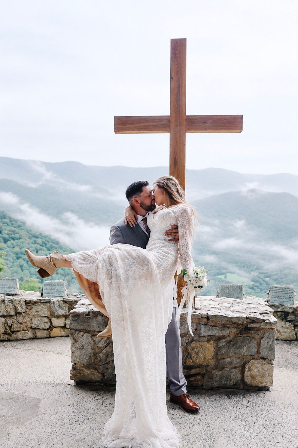 Groom carries bride in lace dress before a wooden cross with misty mountain views at an outdoor wedding ceremony.