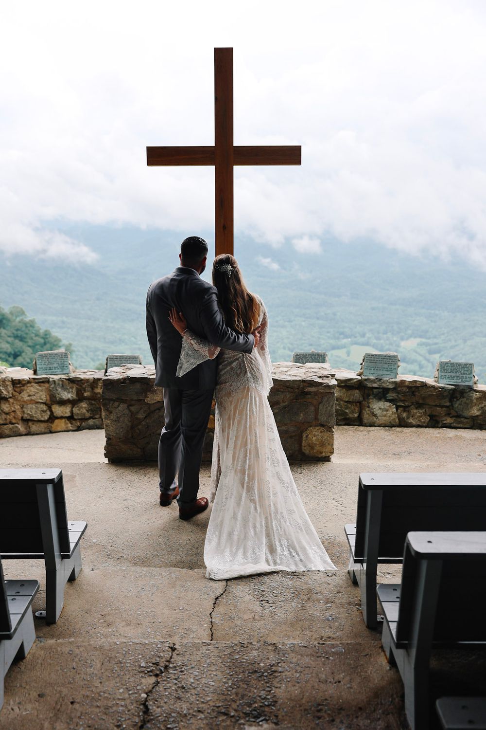 Bride and groom stand before a wooden cross at an outdoor stone chapel with mountain views on their wedding day.