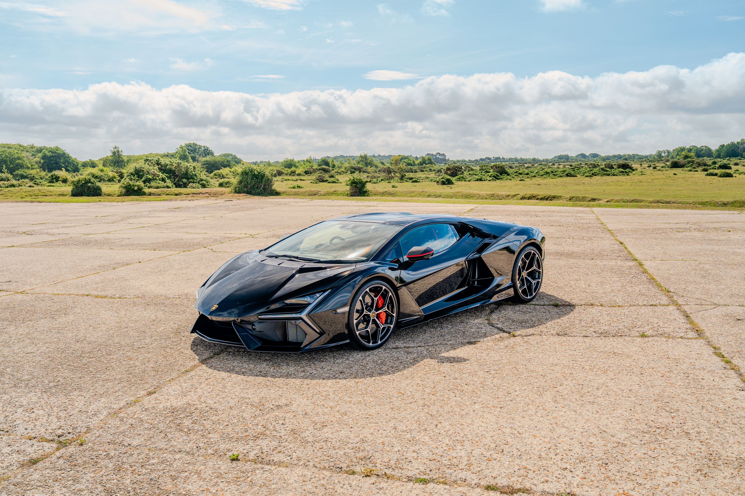 Black Lamborghini supercar parked on open tarmac under cloudy sky, showcasing aggressive angular bodywork.
