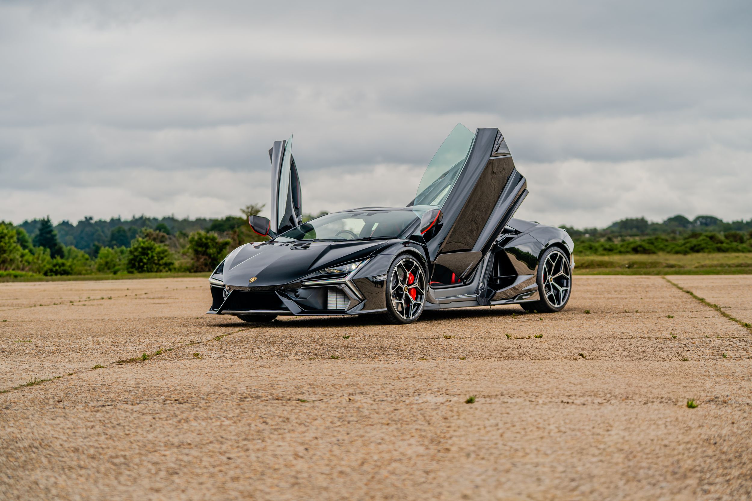 Black Lamborghini supercar with scissor doors open, parked on a tarmac under cloudy skies, showcasing its aggressive design.