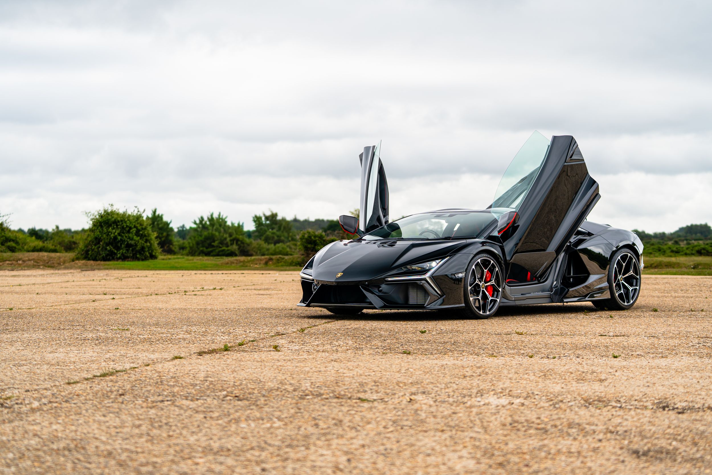 Matte black Lamborghini with scissor doors open, parked on open tarmac under overcast skies.