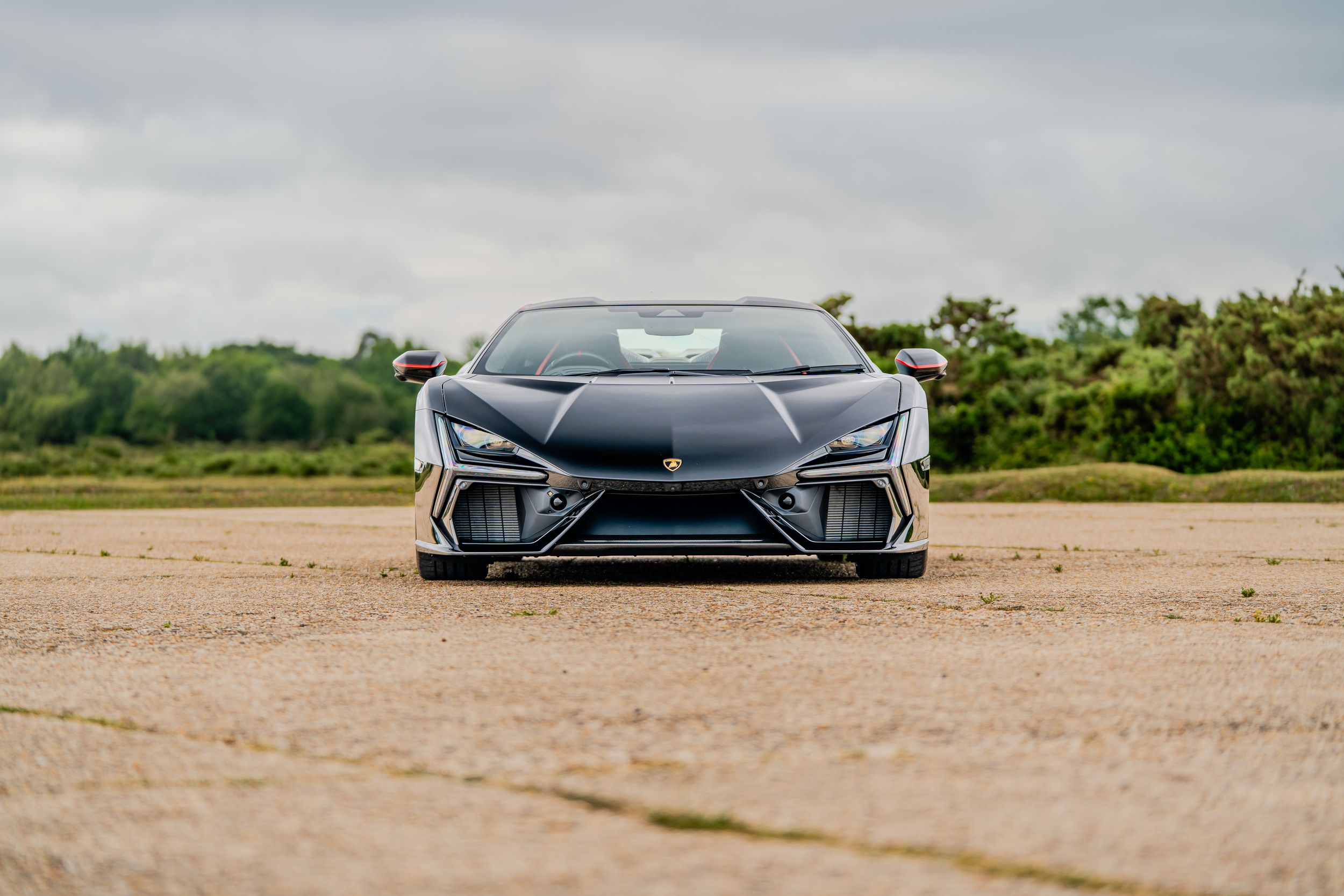 Black Lamborghini supercar front view on open gravel surface with overcast sky and green trees in background.