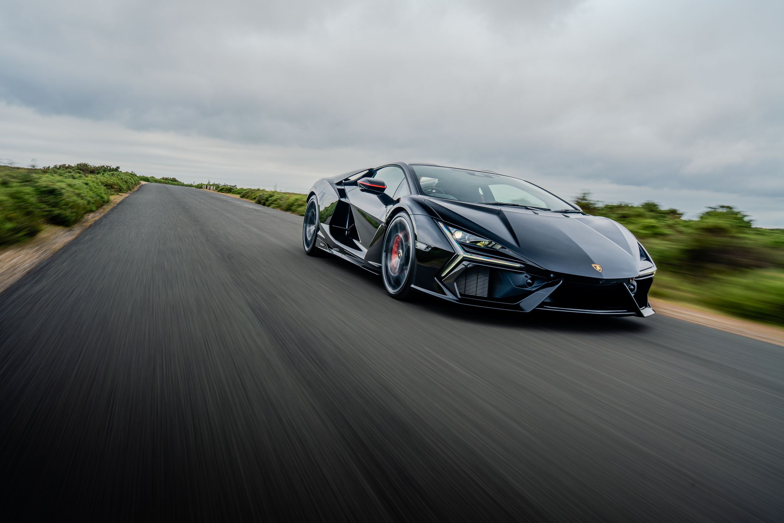Black Lamborghini supercar speeding down an open road with motion blur, captured under a dramatic overcast sky.