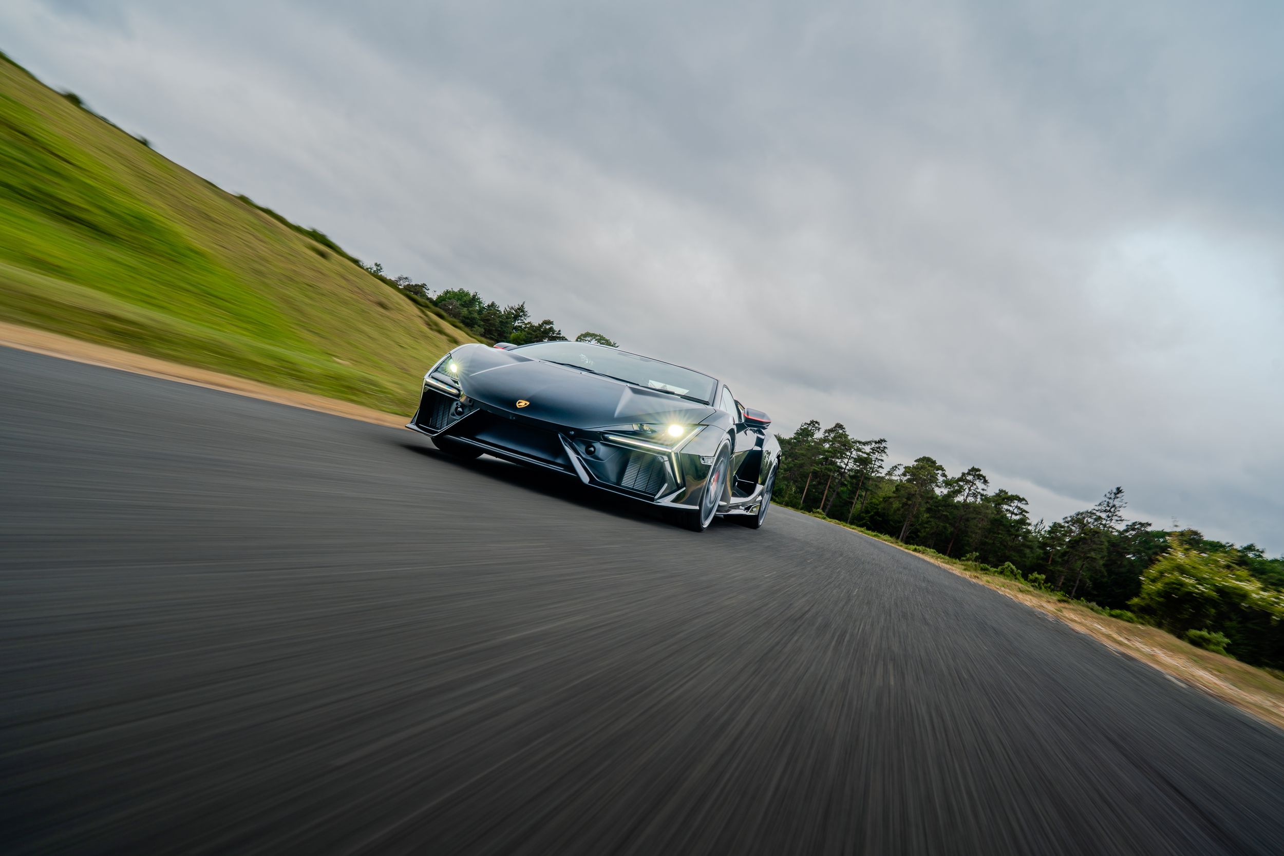 A sleek Lamborghini supercar accelerates on a race track with green hills and cloudy skies in the background.