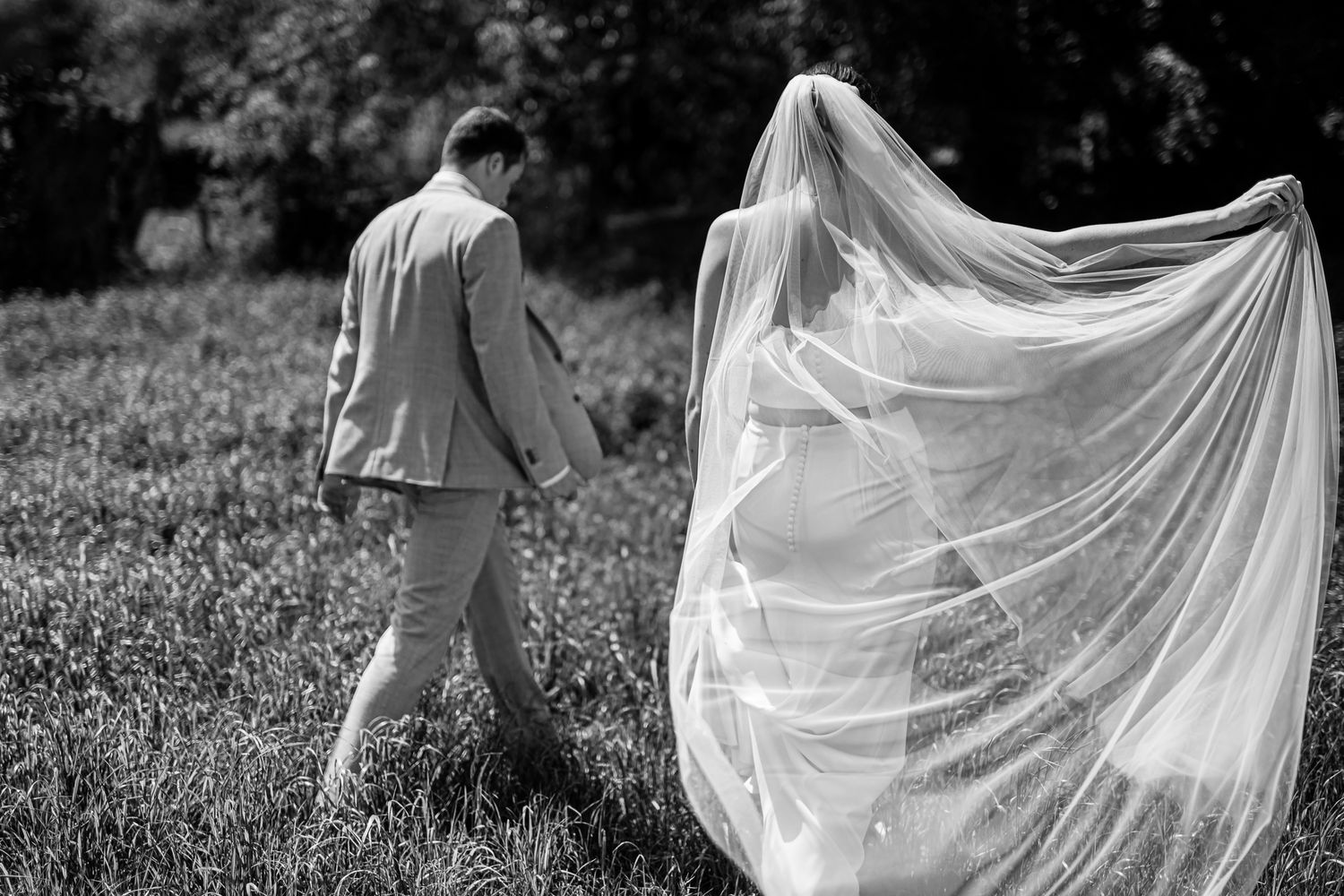 Black and white photo of bride and groom walking in field, wedding veil flowing dramatically in the breeze.