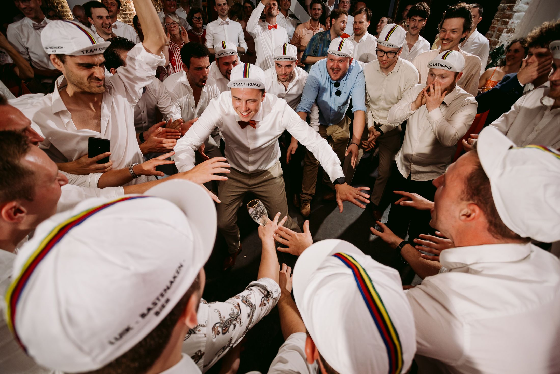 Elderly man in white outfit being celebrated by crowd in matching white caps at a festive cultural gathering.