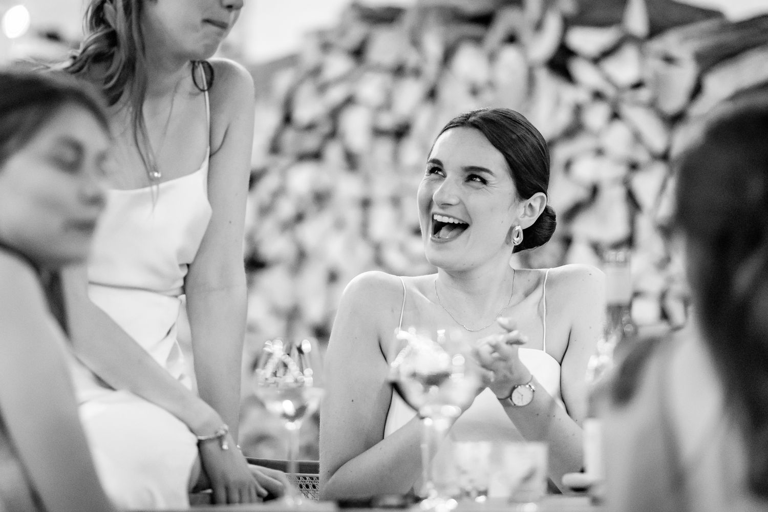 Black and white photo of a laughing woman at a wedding reception table surrounded by guests.