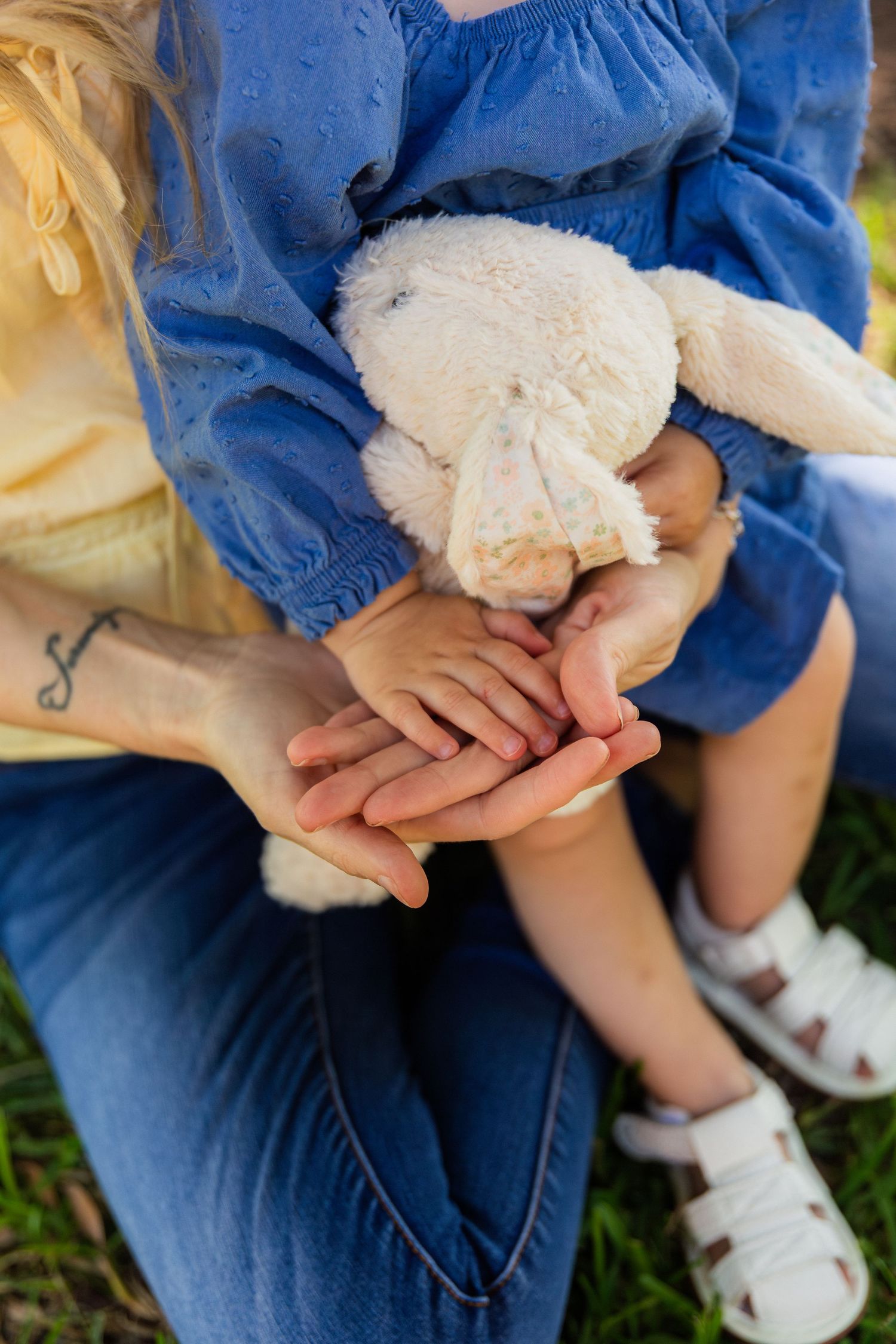 A mother and child holding hands outdoors, with the girl in a blue dress clutching a white stuffed bunny toy.