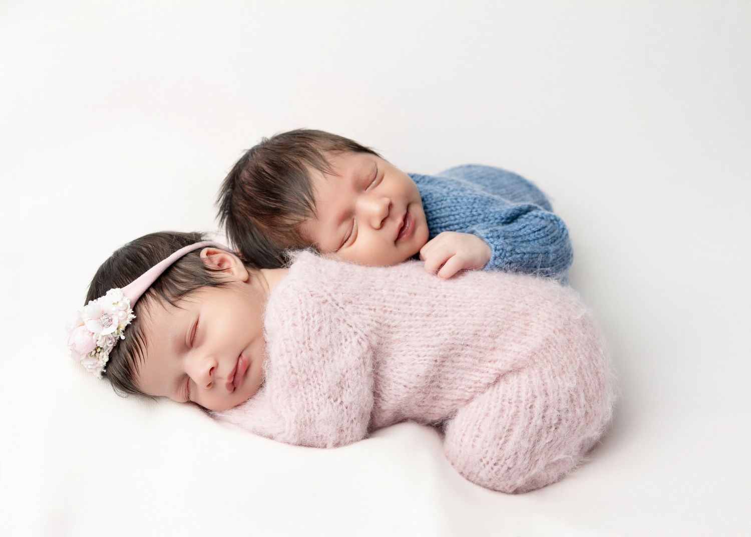 Two sleeping newborn twins wrapped in soft blankets on a white fur rug, one wearing a floral headband.