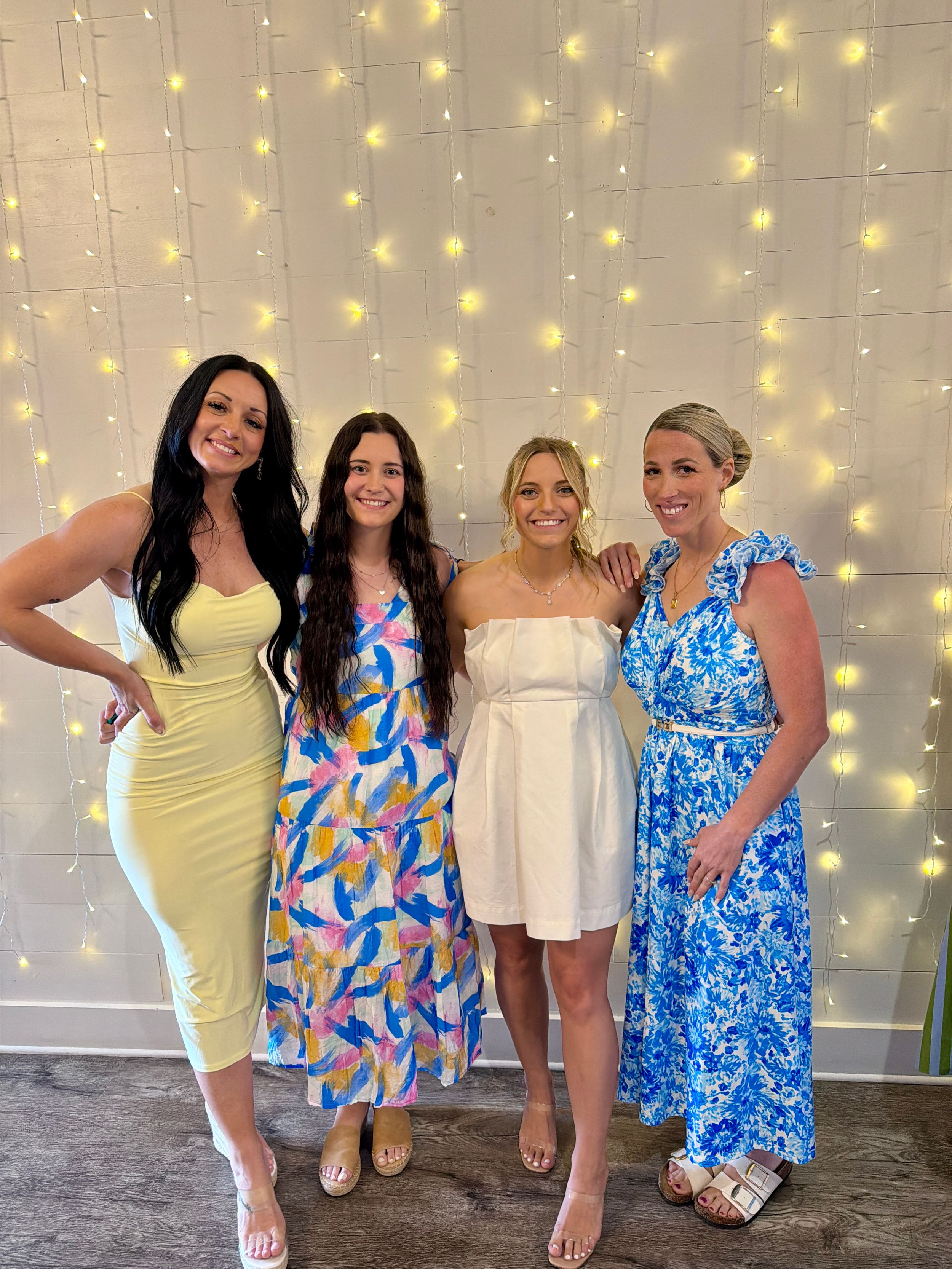 Four women in colorful dresses smile together in front of a twinkling fairy light backdrop at a celebration.