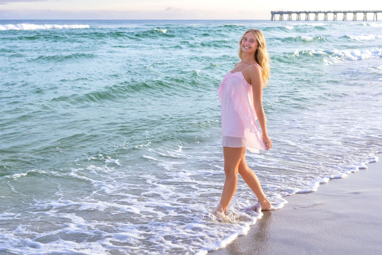 Smiling blonde woman in pink dress walking along sandy beach shoreline with ocean waves and pier in background.