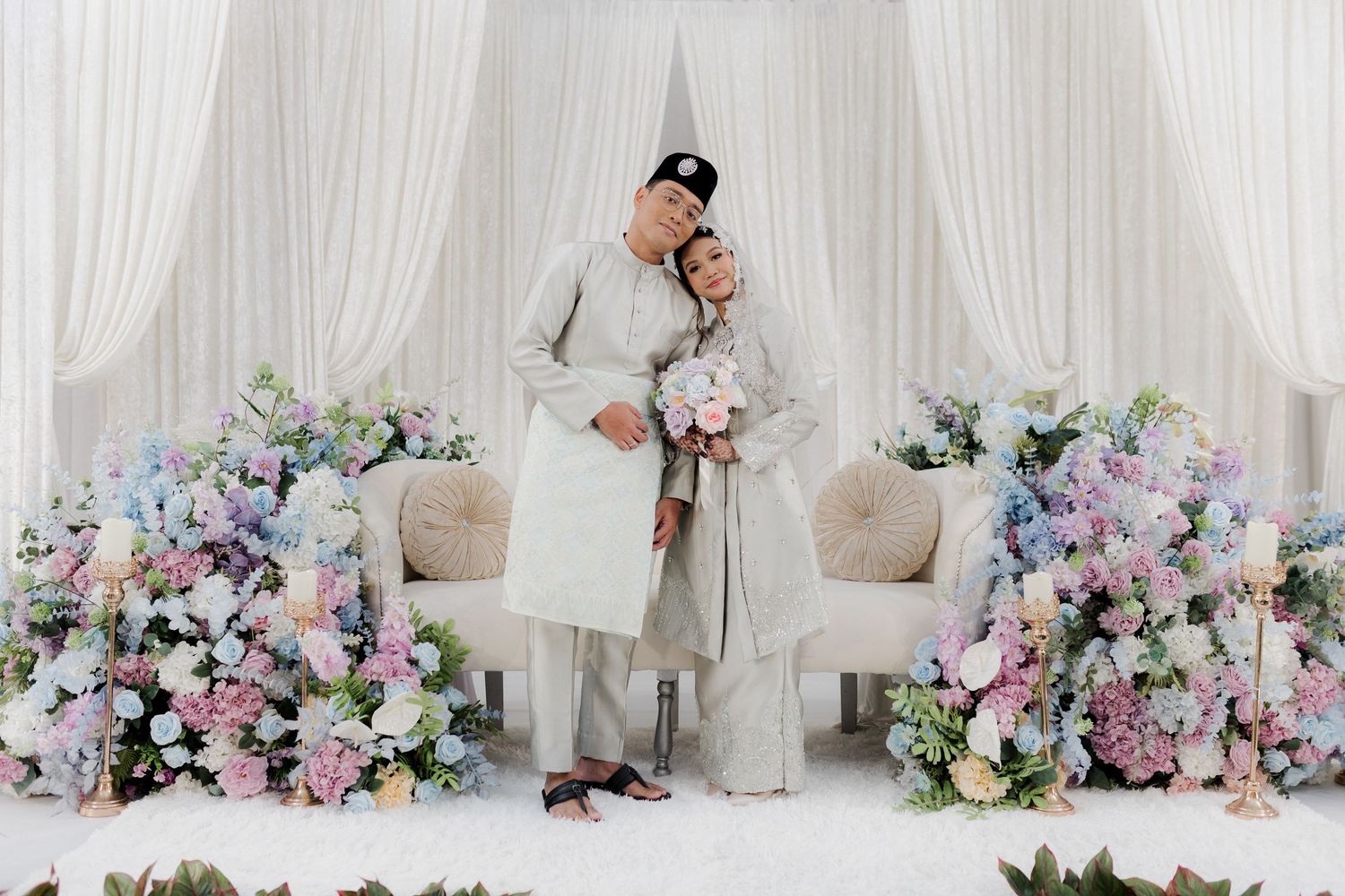 Muslim couple in traditional white attire poses at elegant wedding ceremony with pastel floral arrangements.