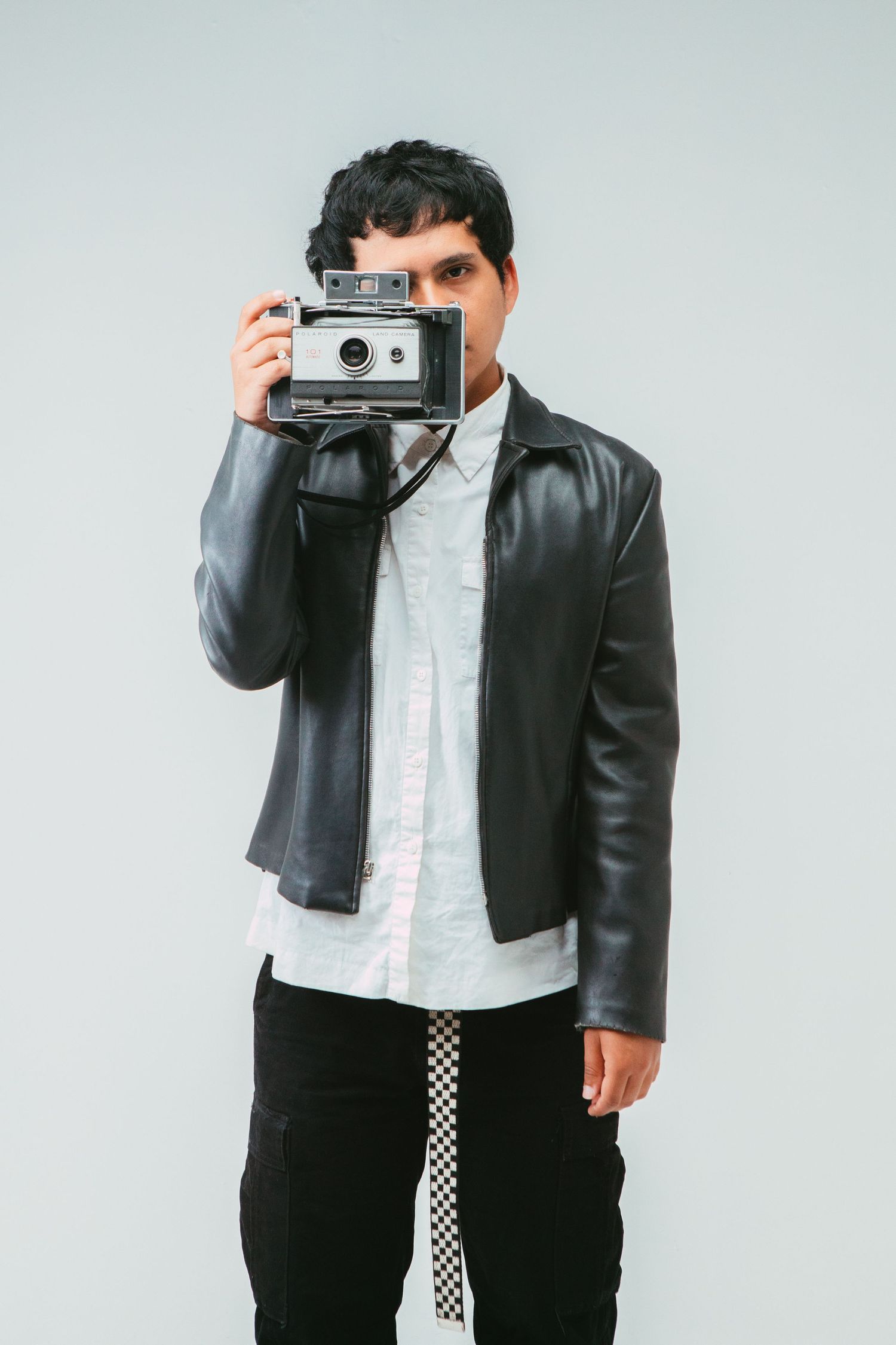A young man in a black leather jacket holds a vintage Polaroid camera up to his face against a light gray background.