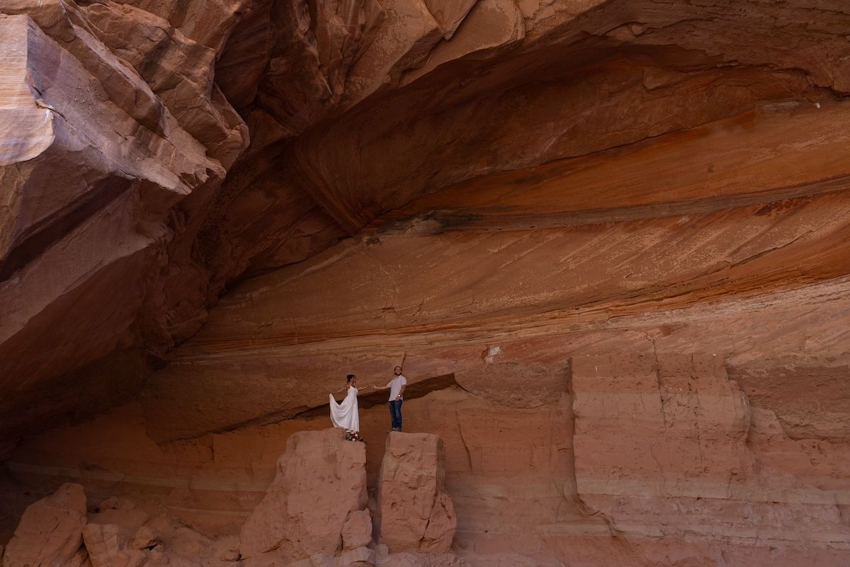 A rock climber scaling a massive red sandstone canyon wall beneath a sweeping natural overhang in a desert landscape.