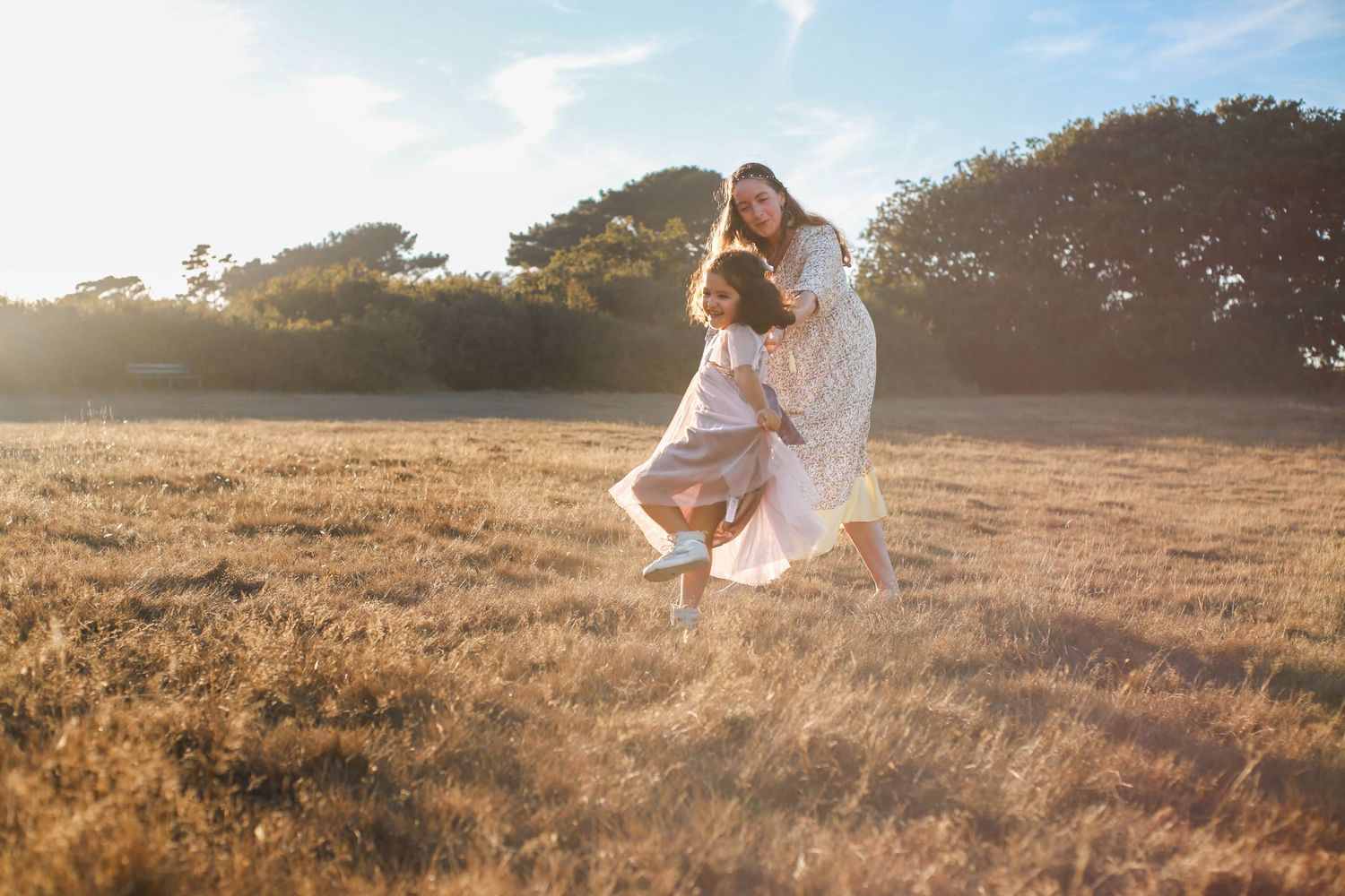 Family playing and lifting child in sunny meadow with tall grass and trees.