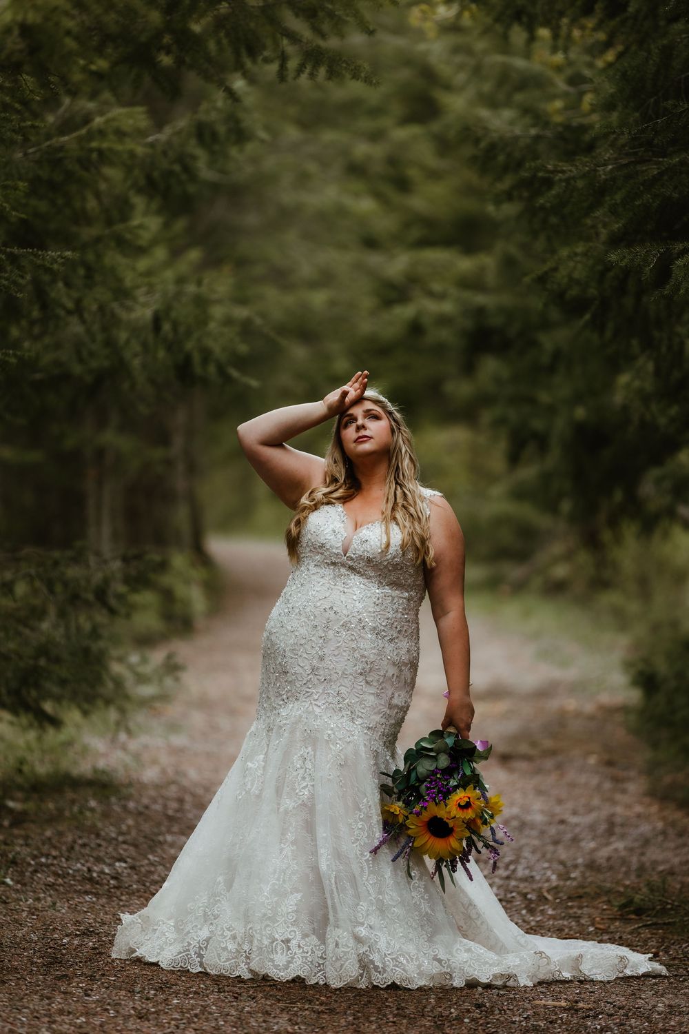 Bride in lace mermaid wedding gown holding sunflower bouquet on forest path, hand shielding eyes from light.