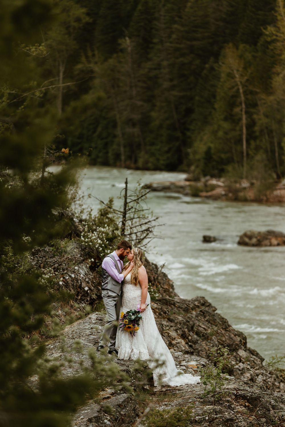Bride and groom kissing on rocky riverbank surrounded by lush forest and rushing turquoise water.