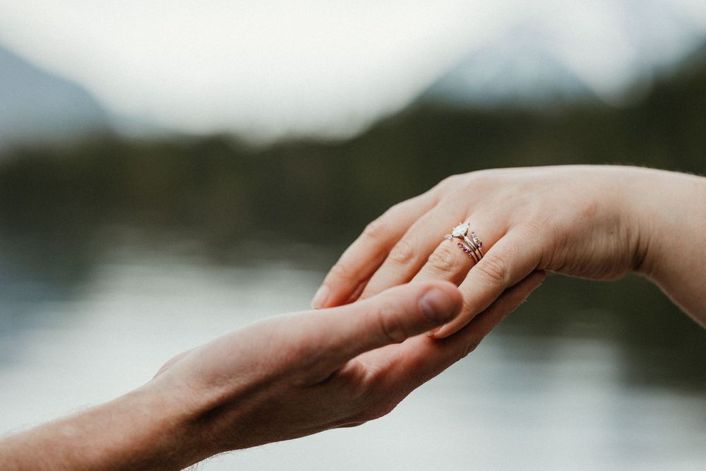 Two hands touching gently, one adorned with a diamond engagement ring, with a scenic mountain lake in the background.