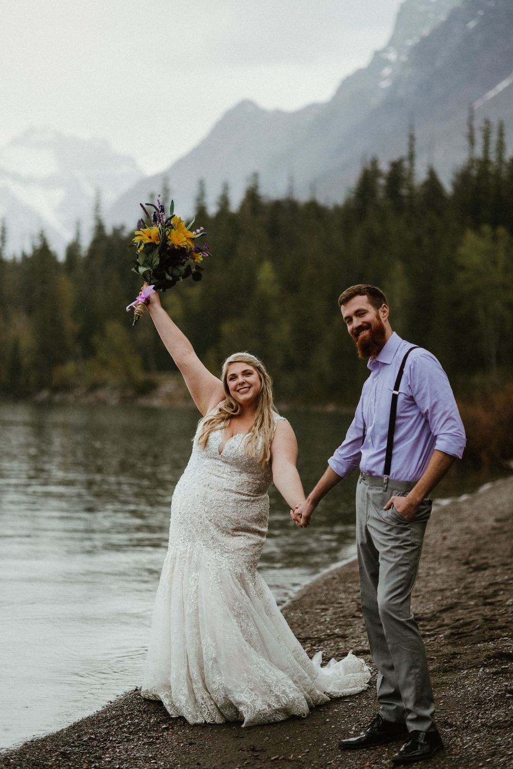 Bride raises bouquet triumphantly beside groom at mountain lake wedding in Banff, wearing lace gown with rustic floral arrangement.