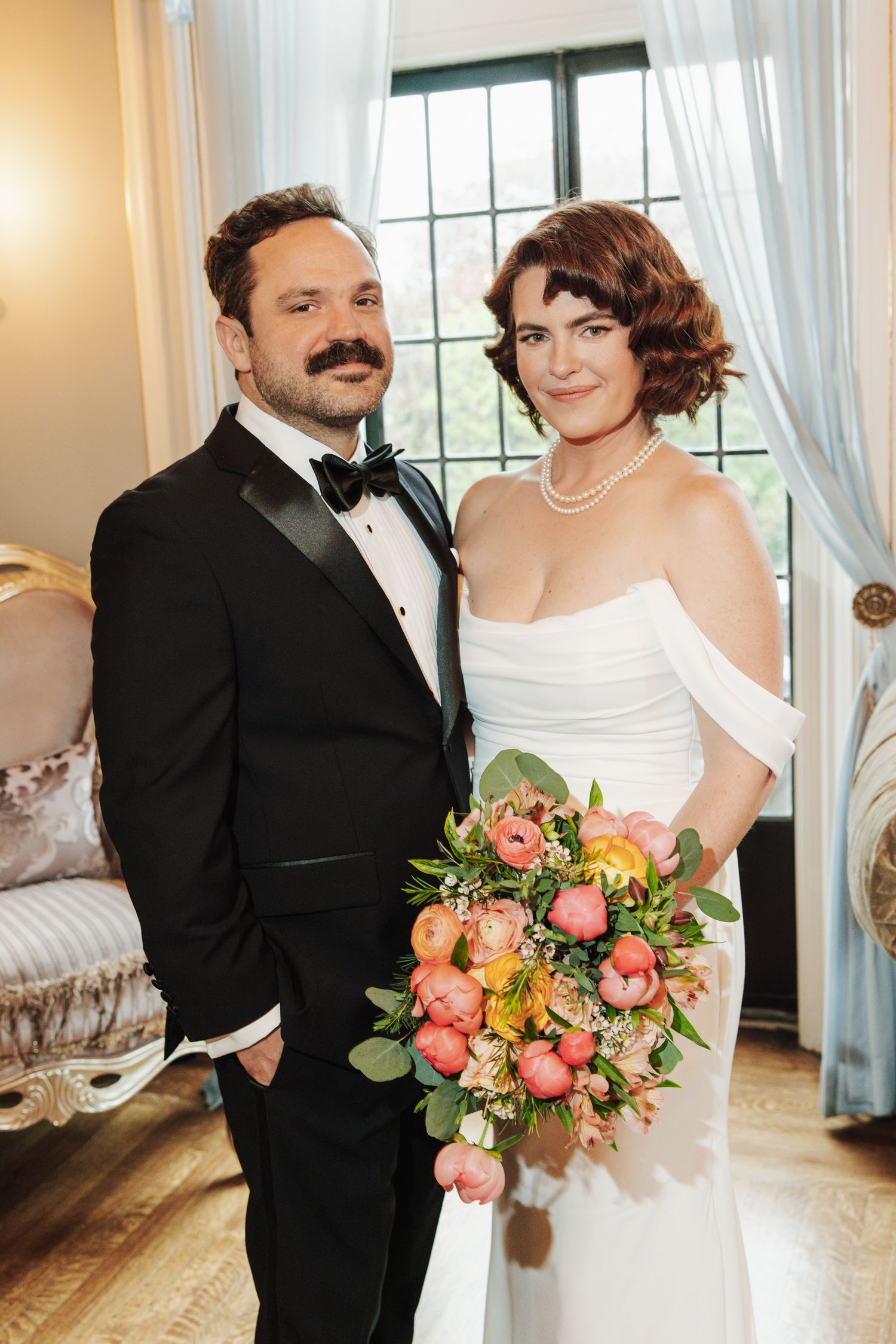 Bride in off-shoulder white gown holding a peony bouquet stands with groom in black tuxedo indoors.