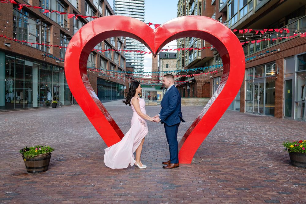 Couple in pink dress and blue suit holding hands inside a large red heart sculpture in a city courtyard.
