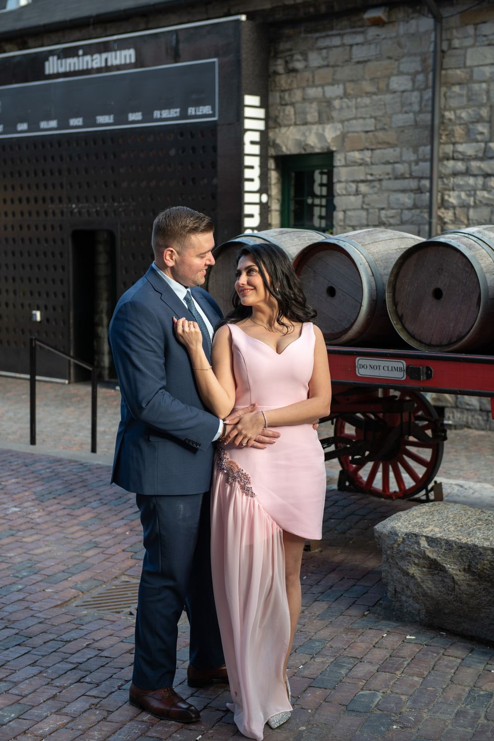 Engaged couple embracing outside Illuminarium venue with wooden barrels and cobblestone street backdrop.