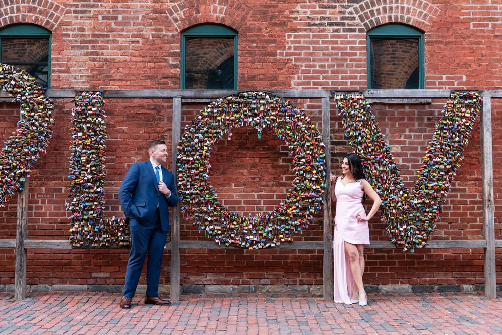 Couple posing apart in front of large LOVE letters made of colorful padlocks on brick wall.