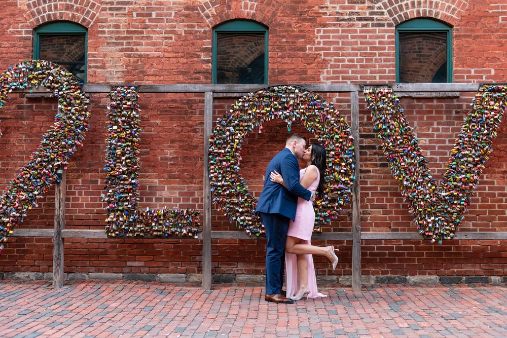 Couple kissing beneath large padlock LOVE letters installation on rustic red brick wall backdrop.