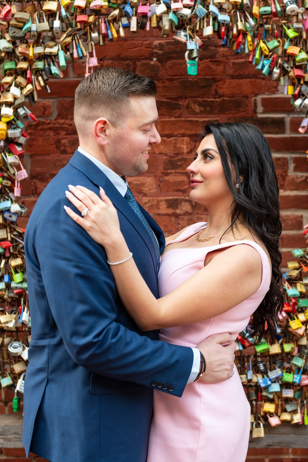 Couple embracing under arch of colorful padlocks on brick wall, gazing into each others eyes.