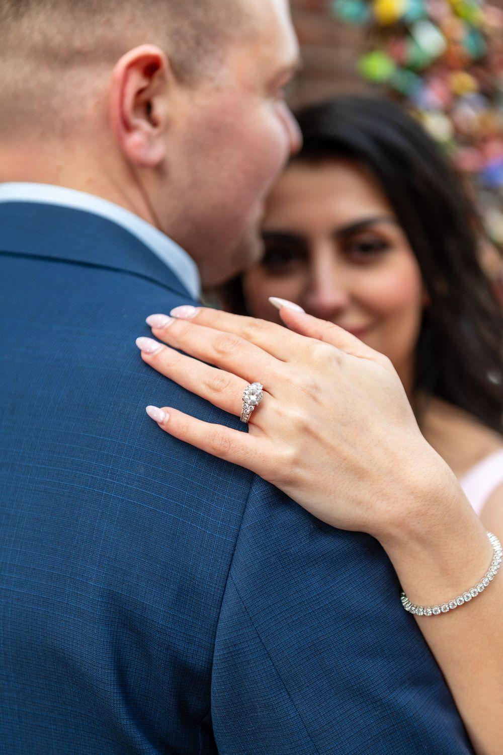 Close-up of diamond engagement ring on womans hand resting on mans navy suit jacket shoulder.