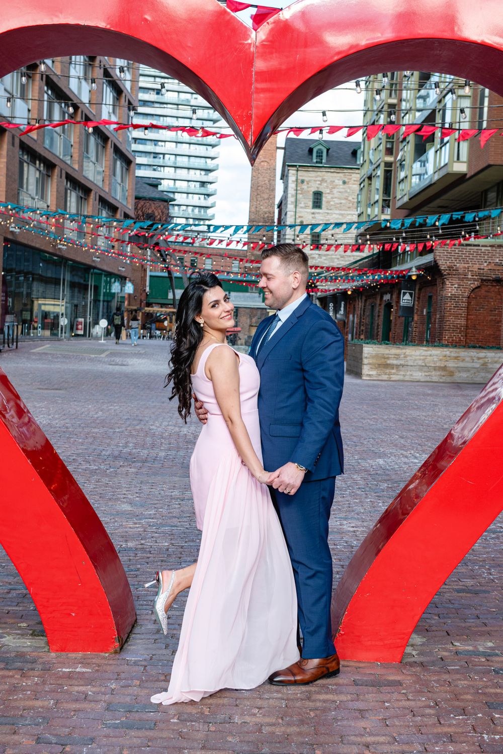 Couple embracing inside a large red heart sculpture, woman in pink gown and man in navy suit in urban setting.