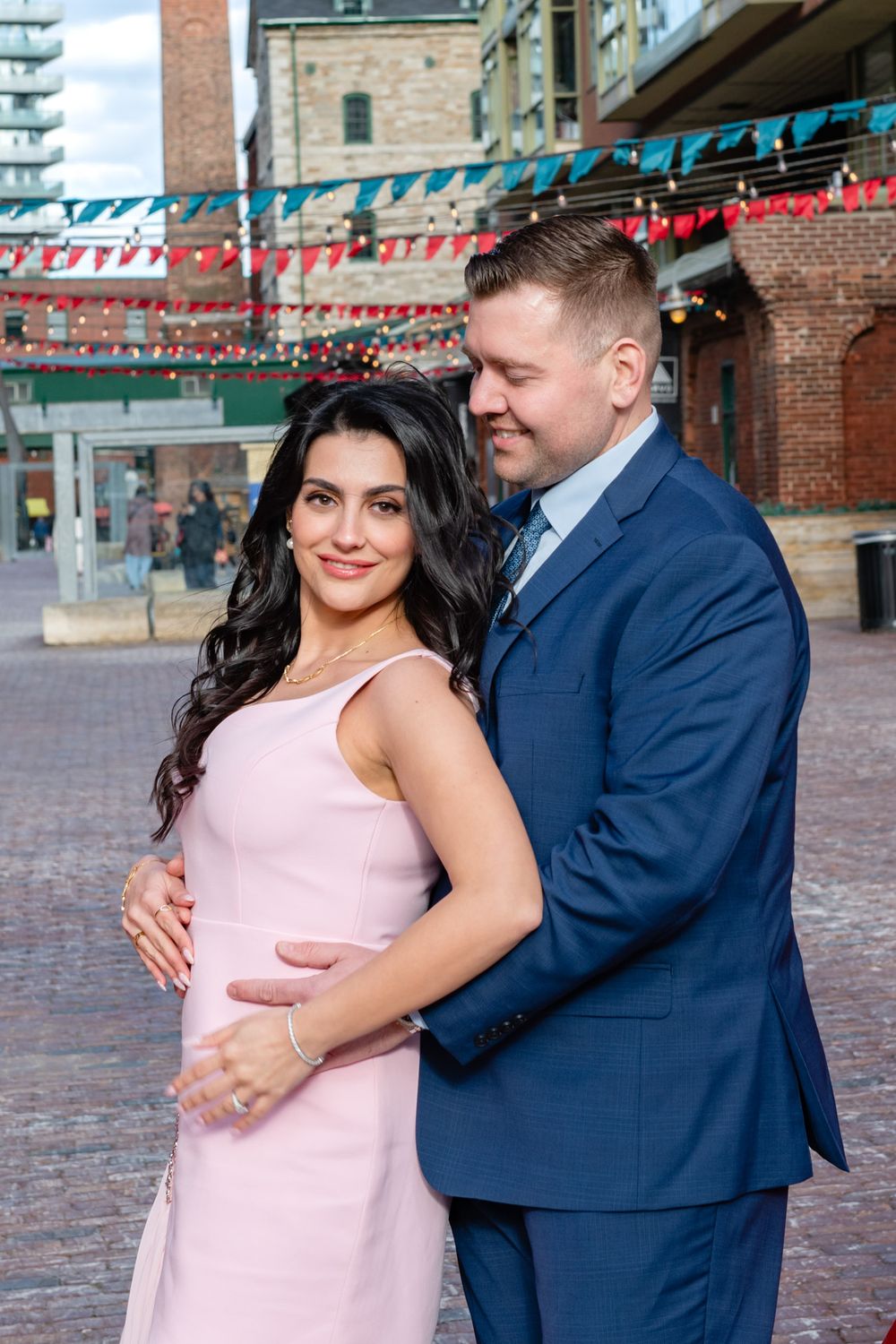 Couple posing in a brick courtyard with colorful bunting flags, woman in pink dress embraced by man in navy suit.