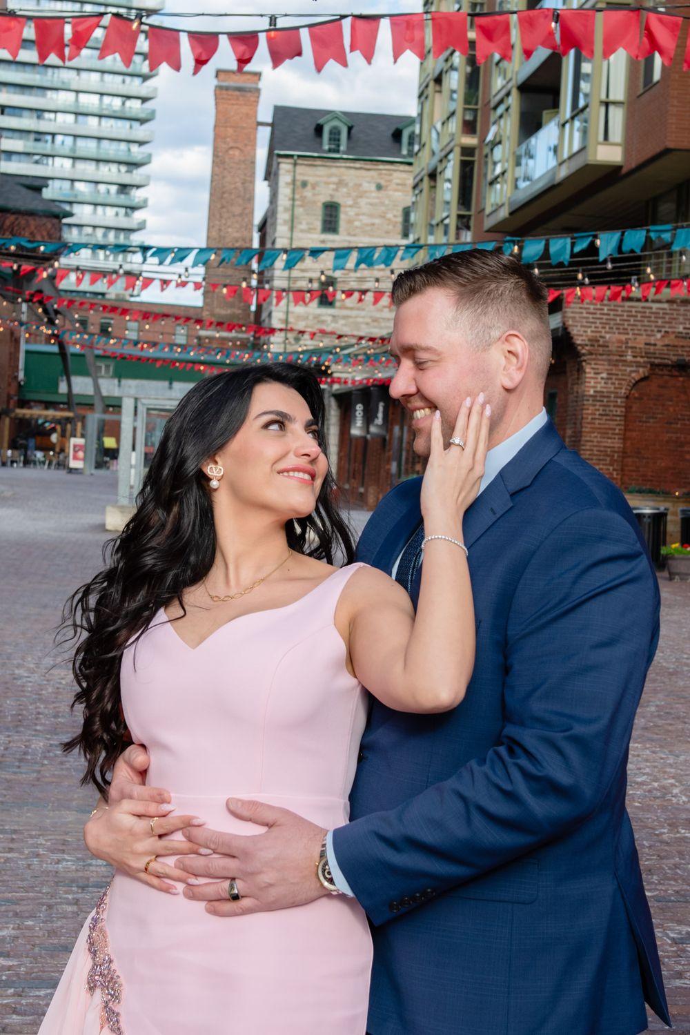 Couple sharing a tender moment in a brick courtyard with colorful bunting, woman in pink dress touches mans face.