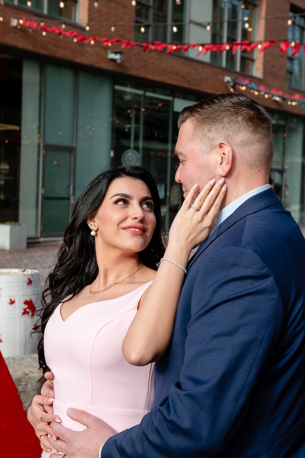 Couple sharing a romantic moment outdoors, woman in pink dress touching mans face, string lights in background.