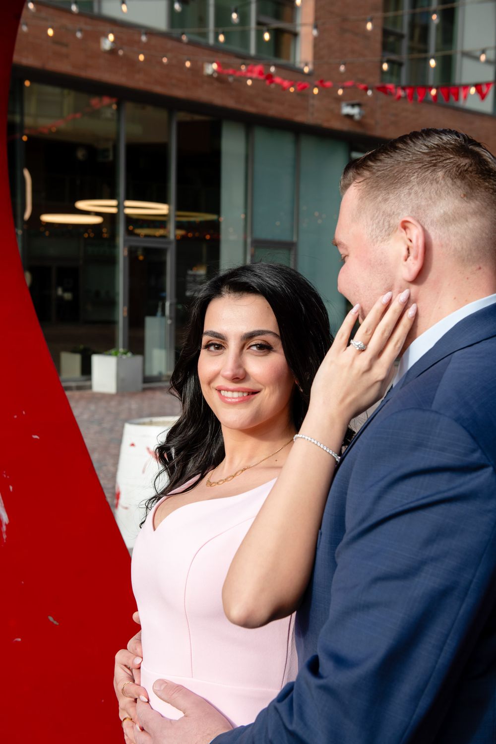 Engaged couple embracing near red heart sculpture, woman in pink dress smiling at camera, urban setting.
