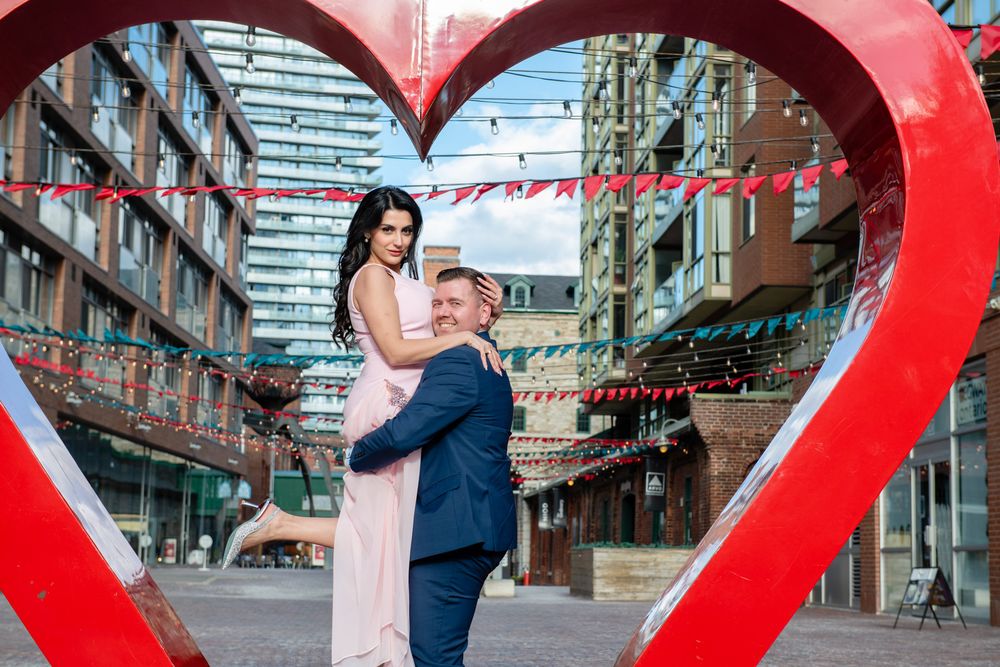 Man lifting woman in pink dress inside red heart sculpture, romantic engagement photo in city district.