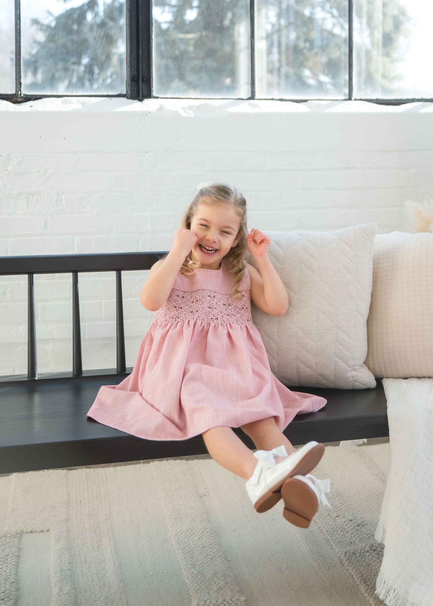 A joyful little girl in a pink smocked dress and white shoes sits laughing on a bench indoors.