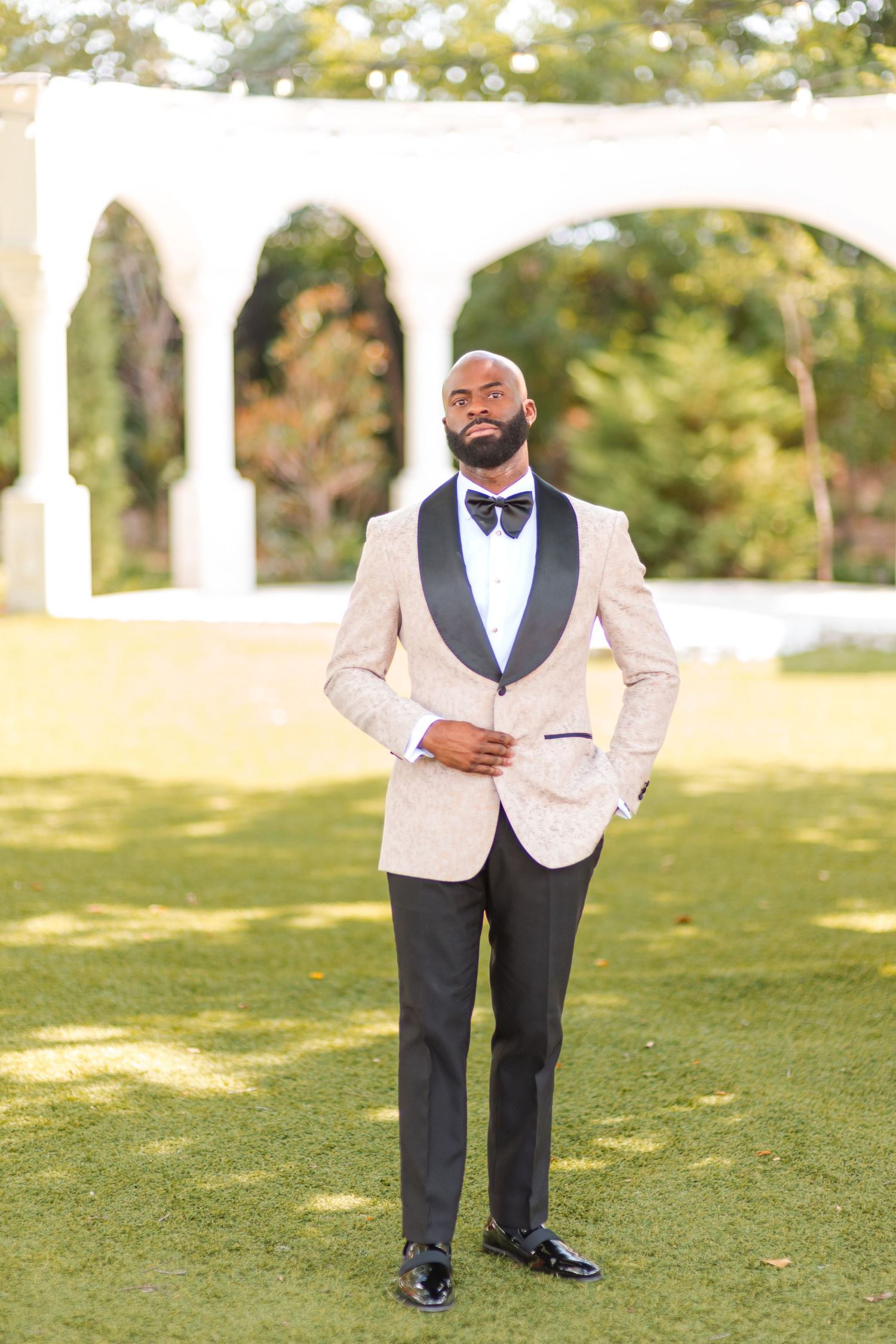 Elegant groom wearing cream tuxedo jacket with black lapels and bow tie poses outdoors by white archway.