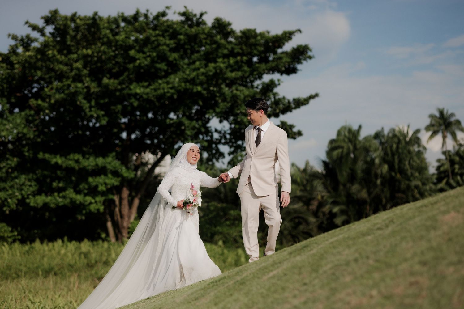 Muslim bride in white hijab and gown holding hands with groom in cream suit walking on green hillside.