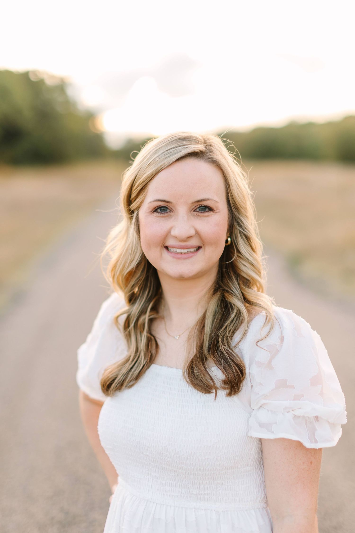 A smiling blonde woman in a white smocked dress poses on a sunlit dirt road surrounded by golden fields.