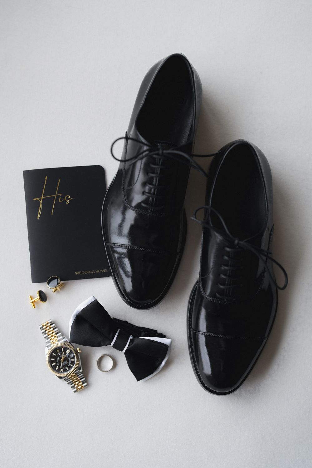 Black oxford dress shoes with bow tie, cufflinks, watch, ring, and wedding vows booklet arranged for a groom.