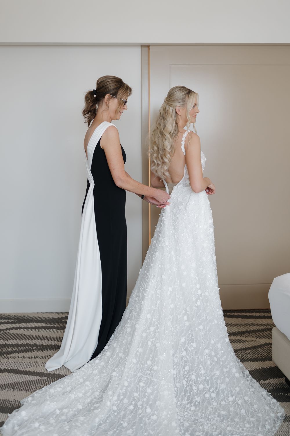 Bride in floral lace gown being helped by woman in black and white dress during wedding preparation.