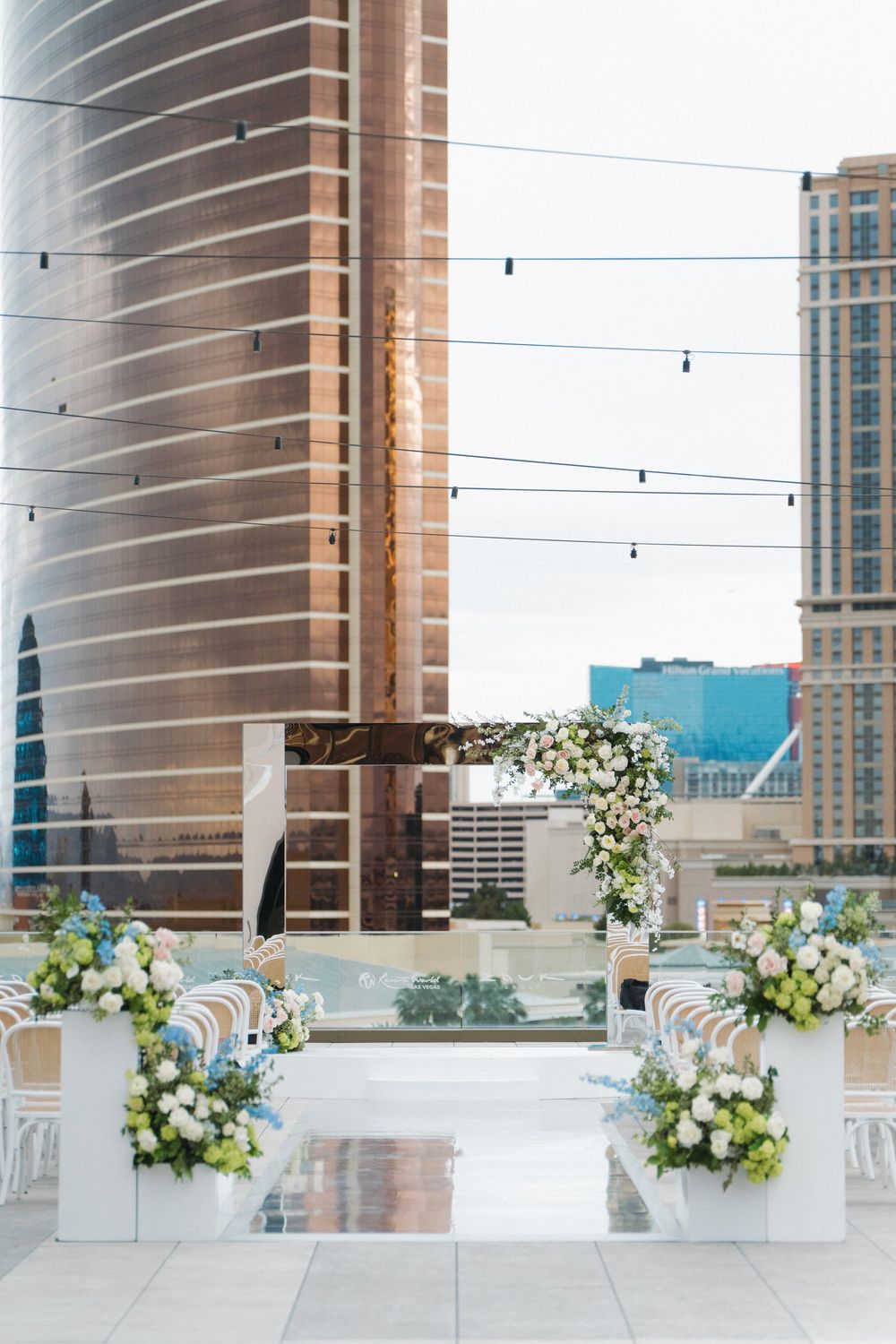 Rooftop wedding ceremony setup with white floral arrangements and mirrored aisle against a city skyline backdrop.