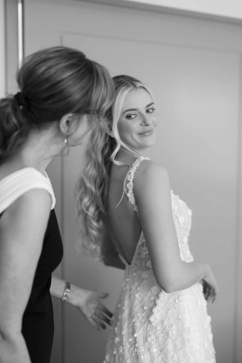 Black and white photo of bride in lace gown smiling at mirror while woman helps fasten her dress.