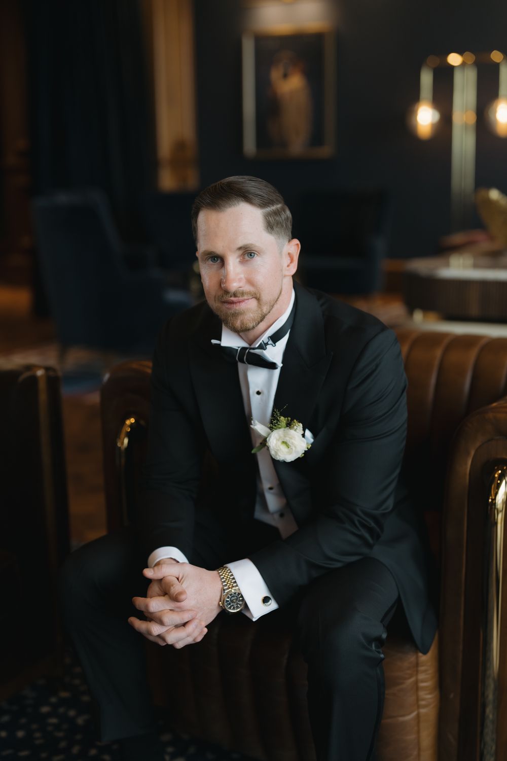 Dapper groom in black tuxedo with bow tie and boutonniere sits in leather chair before wedding.