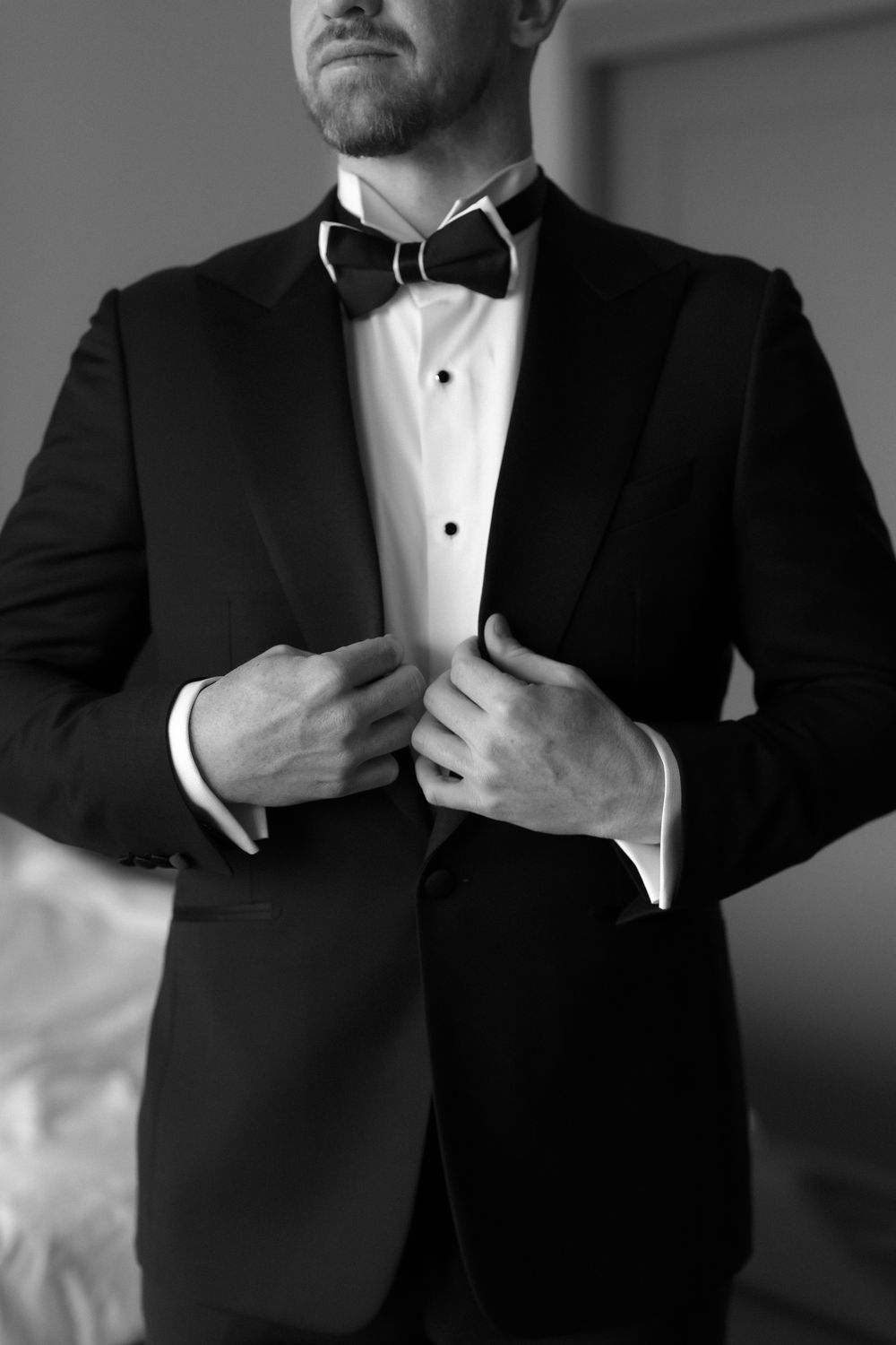 Black and white photo of a groom in a classic tuxedo adjusting his bow tie before the wedding ceremony.