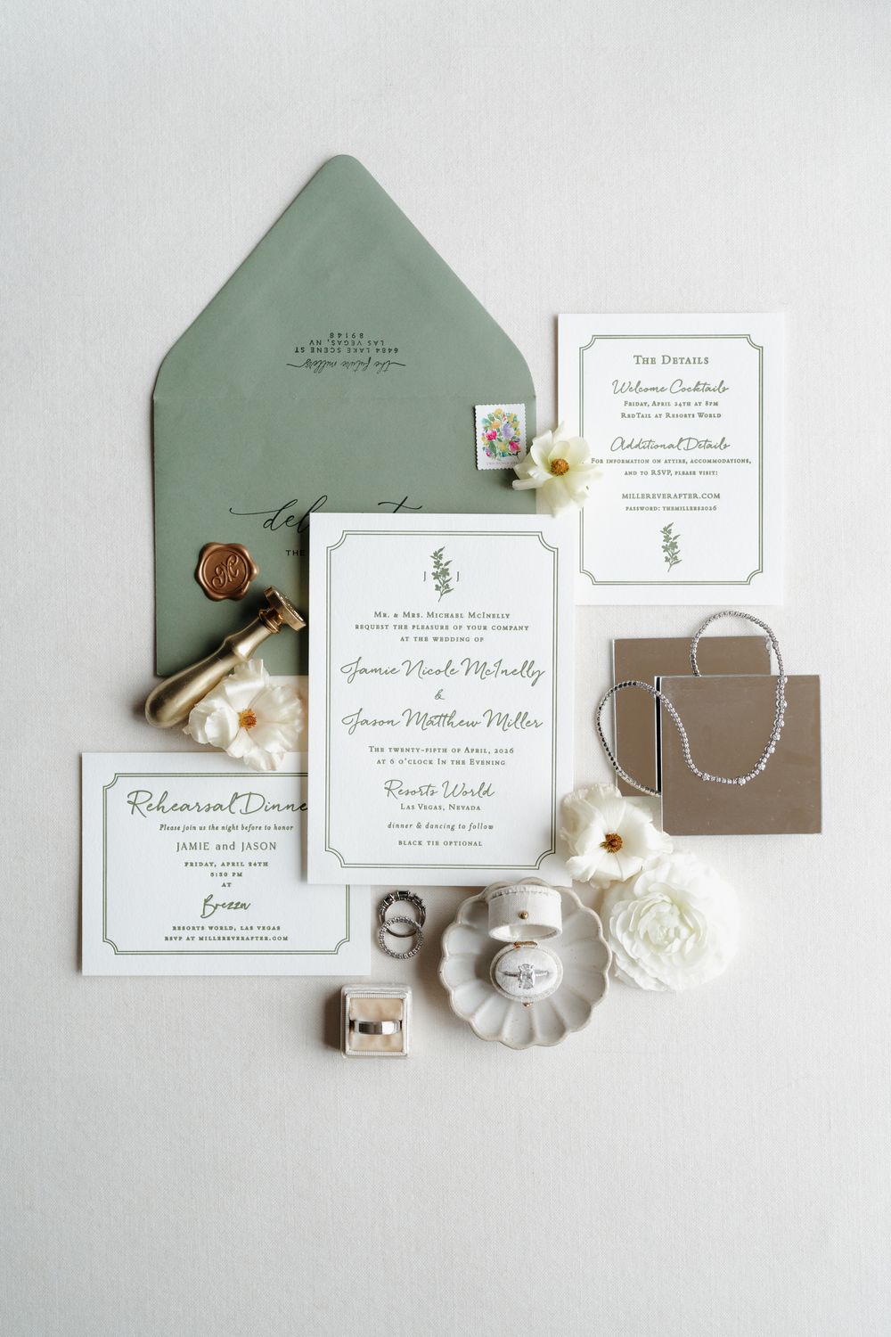 Elegant wedding invitation suite with sage green envelope, wax seal, white florals, and jewelry on a white background.