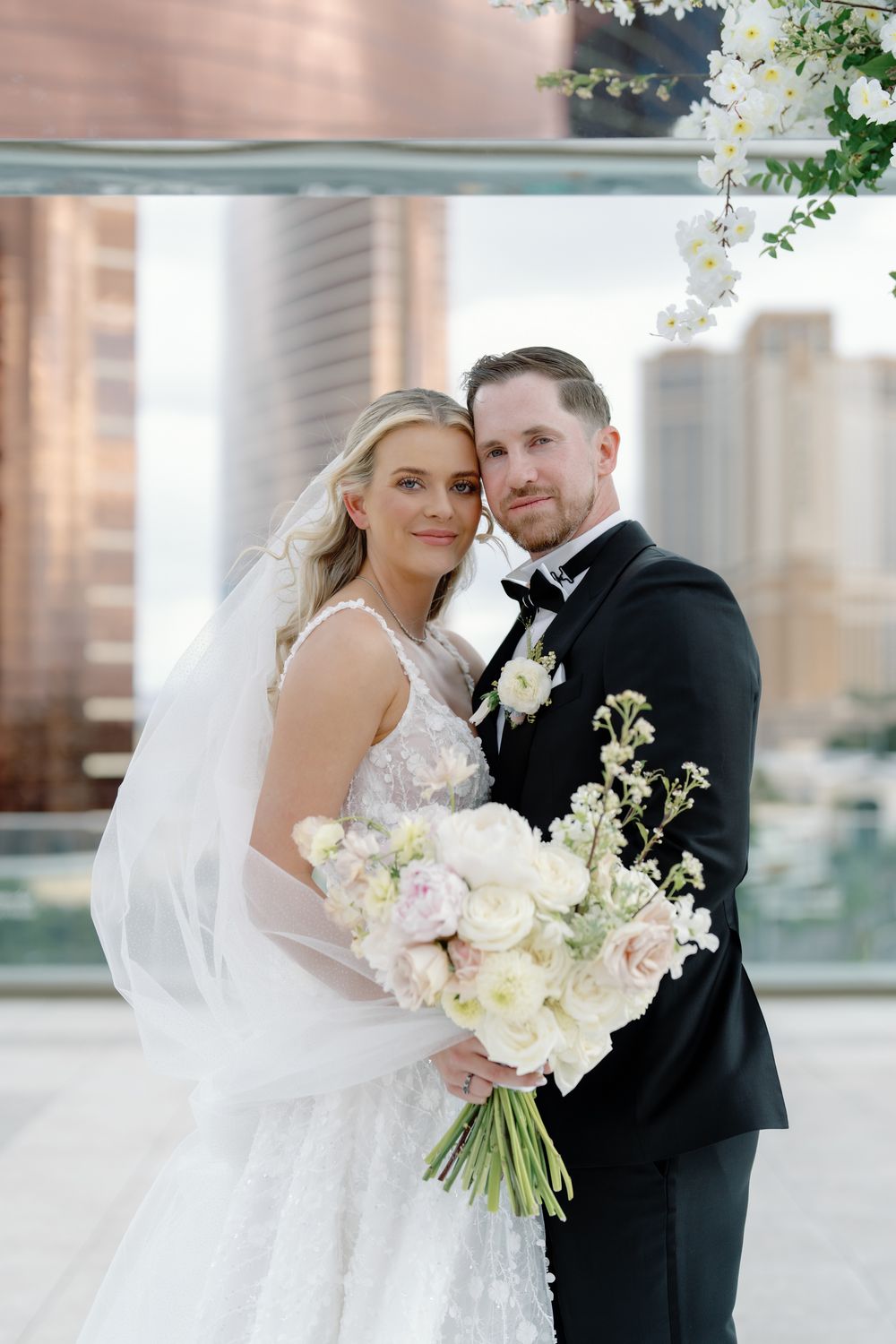 Bride in floral lace gown with veil and groom in black tuxedo posing on rooftop with city skyline.