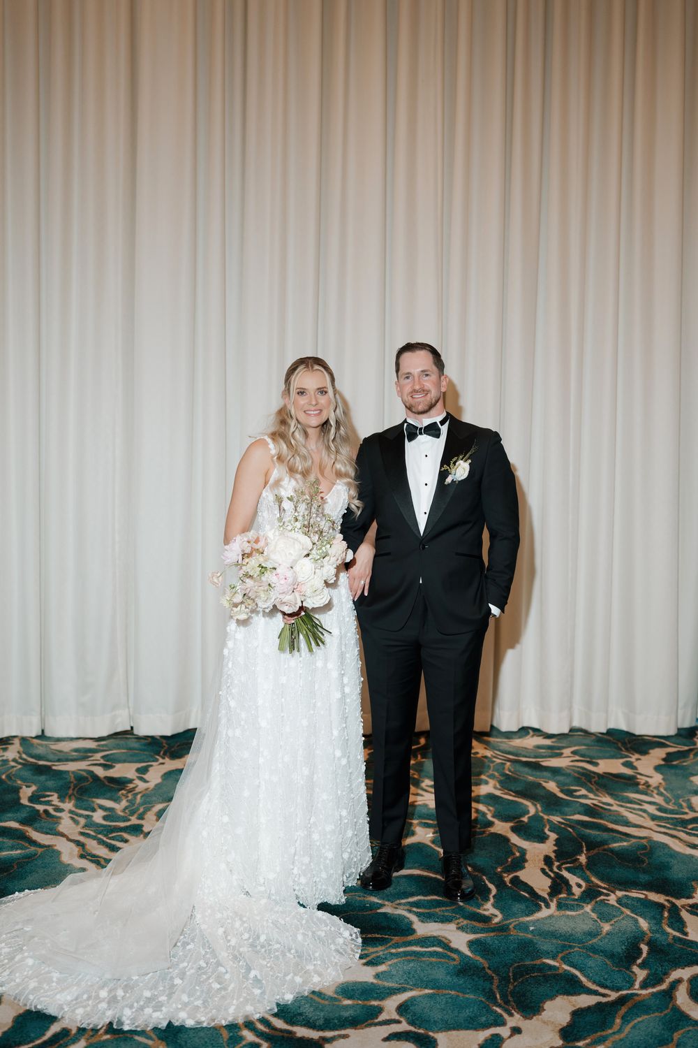Bride in floral lace gown holding pink bouquet stands with groom in black tuxedo before ivory drape backdrop.