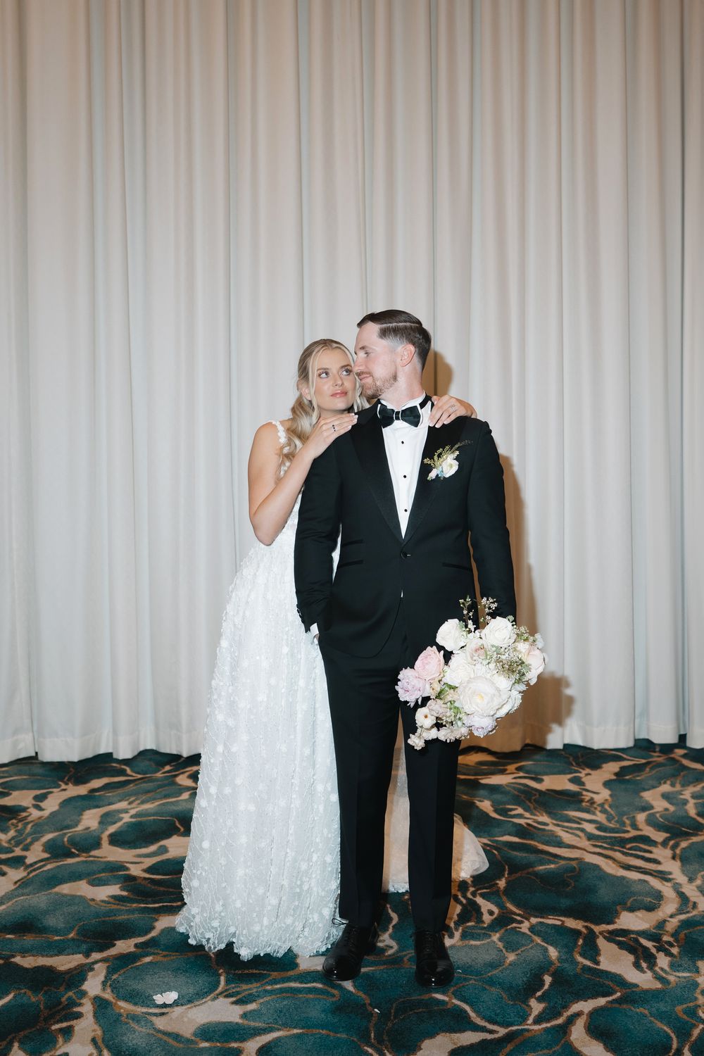 Bride in white floral gown embraces groom in black tuxedo holding pink bouquet against draped backdrop.